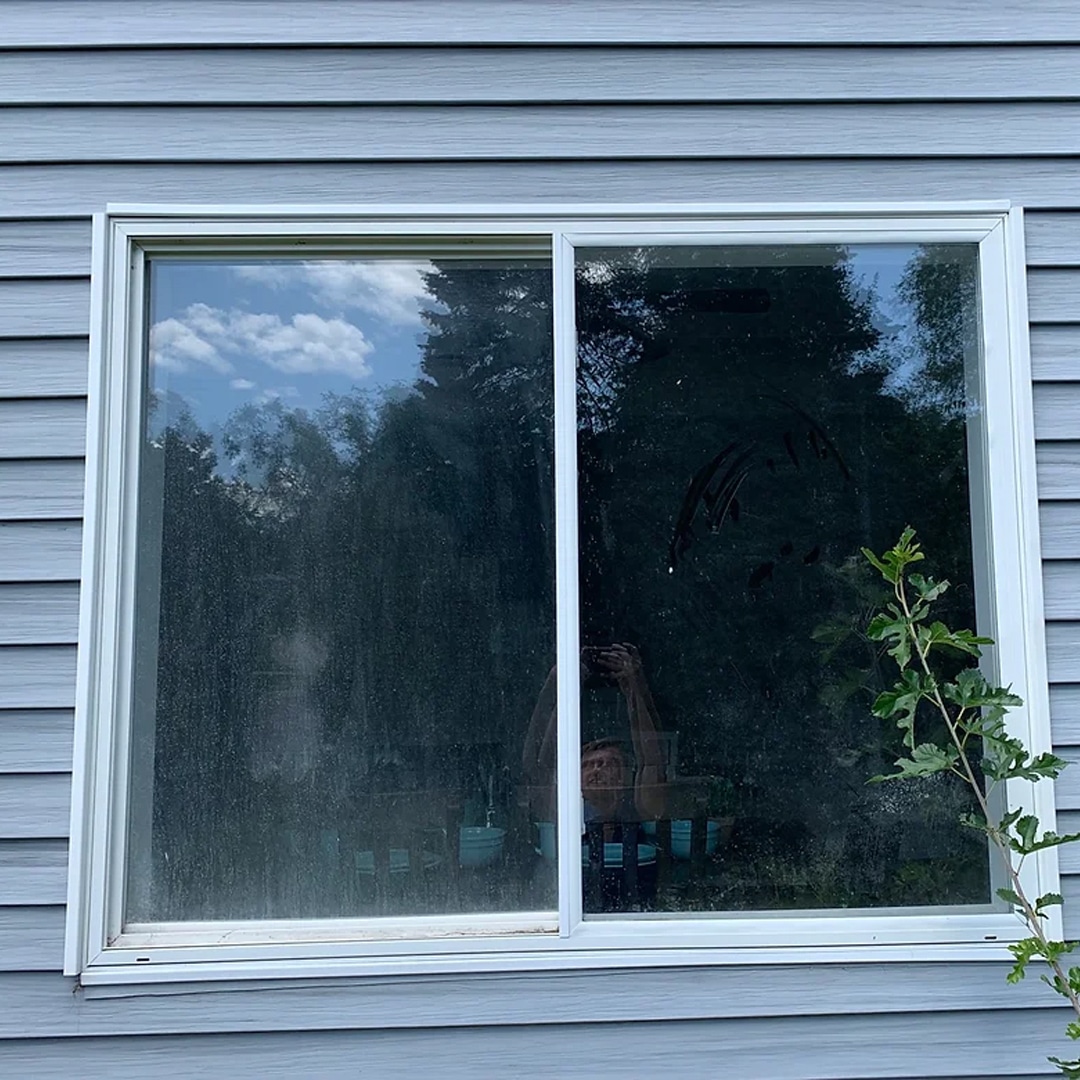 A sliding window on a gray siding wall with reflections of trees and sky. There is a faint drawing on the dirty glass—an ideal spot to showcase the results of professional exterior cleaning services. A green plant grows nearby.