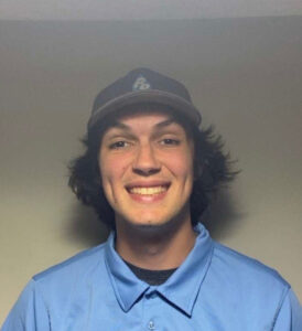 A young man with medium-length dark hair, wearing a blue collared shirt and a baseball cap, smiles at the camera against a plain background—meet-purepro-team.