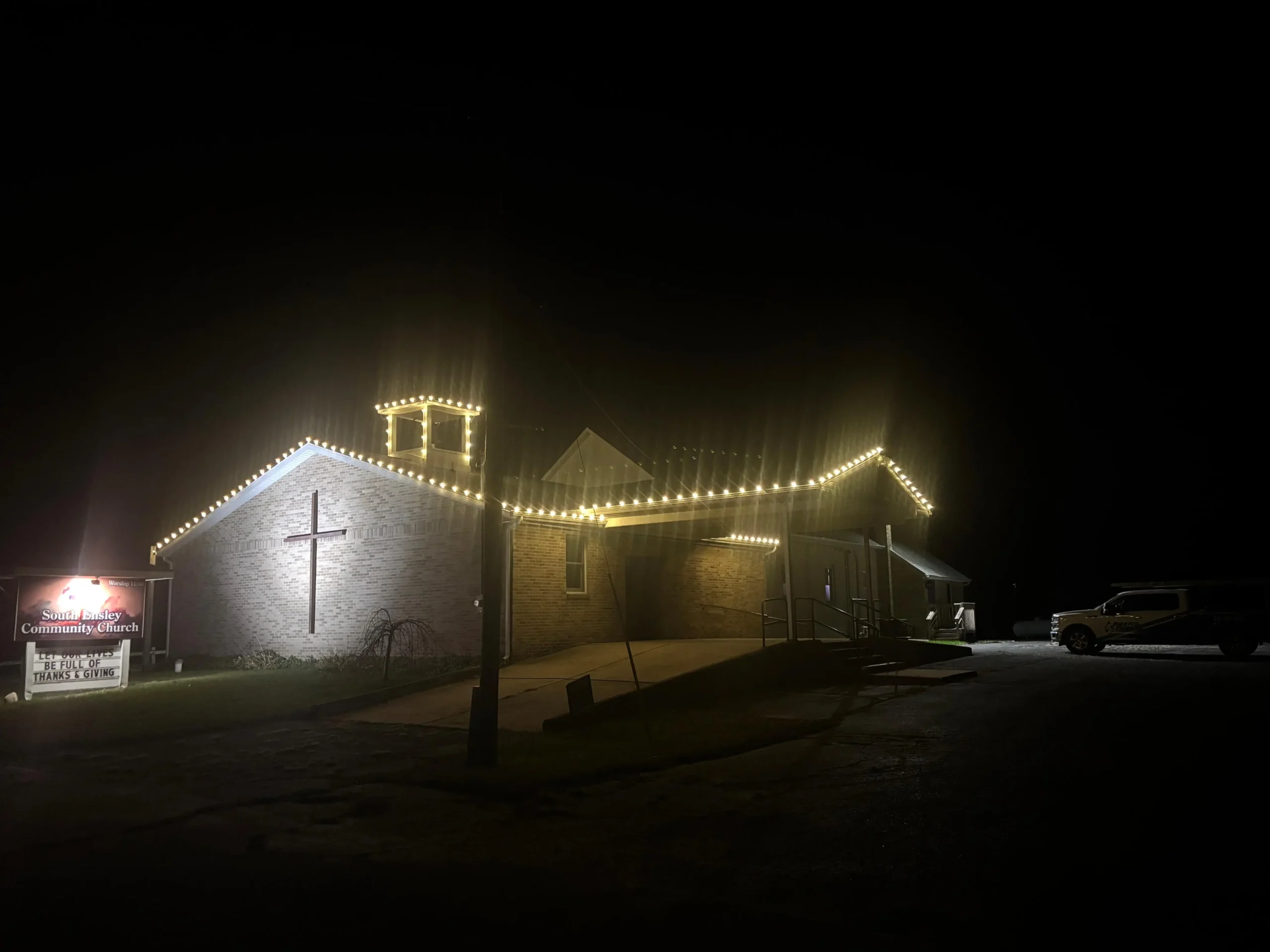 A church building at night with exterior string lights illuminated, a lit cross on the wall, a signboard out front, and a parked vehicle to the right.