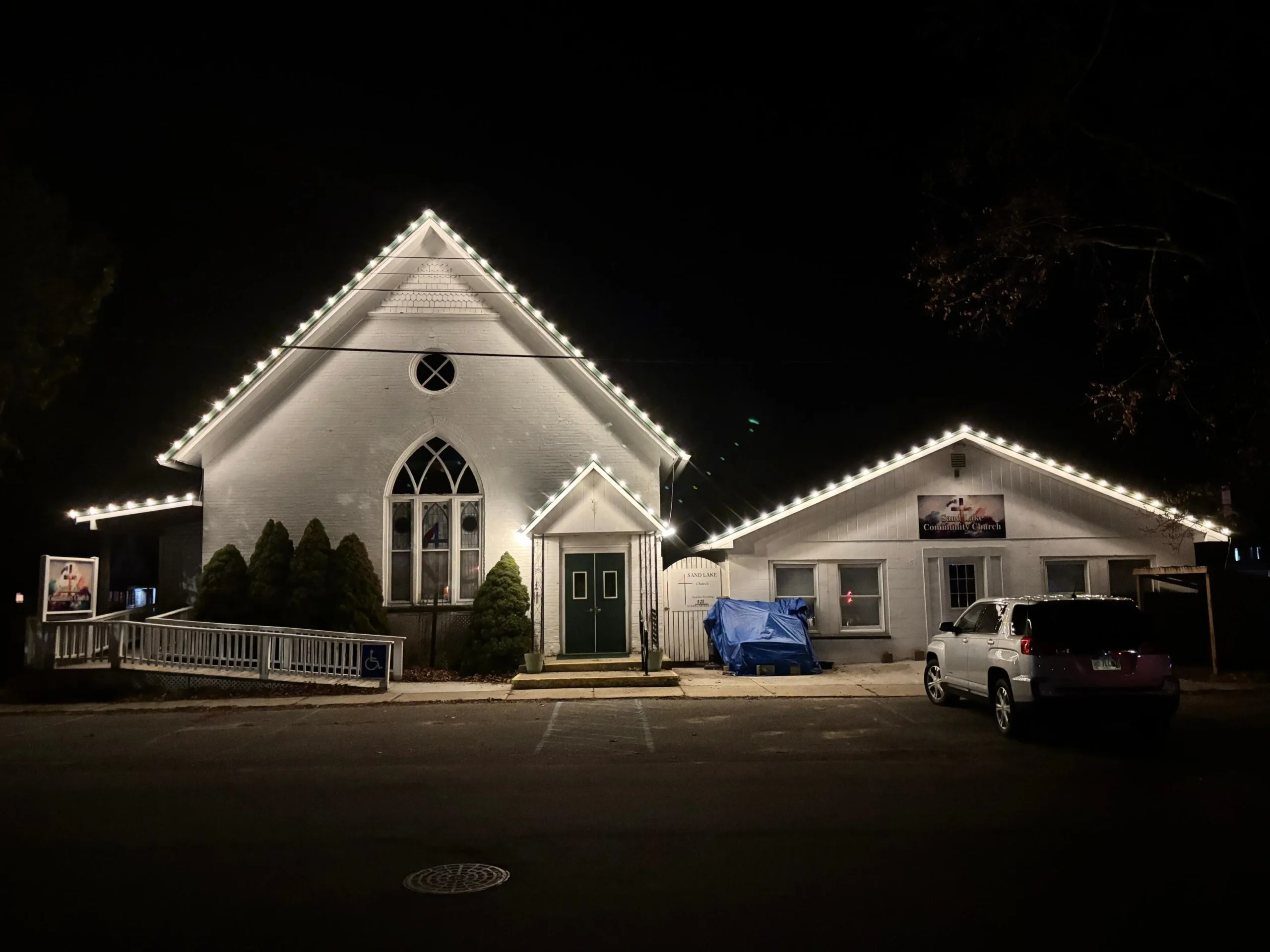 A white building with peaked roofs outlined in string lights at night, featuring a main entrance, ramp, parked car, and covered object in front.