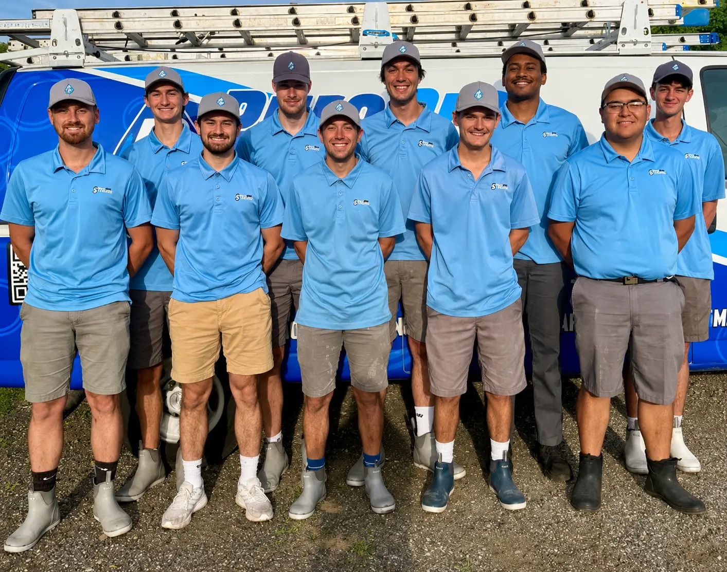 Ten men wearing light blue polo shirts, shorts, and boots stand in two rows in front of a blue service van with a ladder on top.